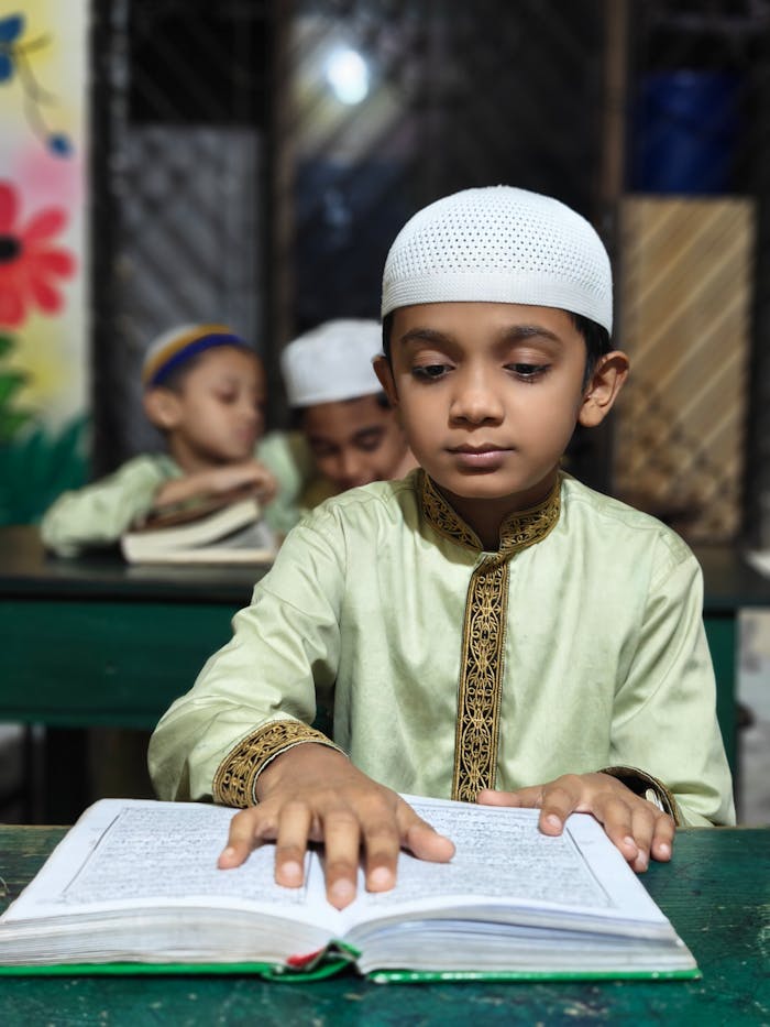 A young boy in a traditional outfit reading the Quran in a classroom setting.